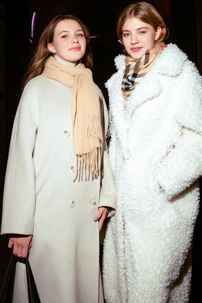 Two women in stylish warm coats and scarves, posing outdoors on a cool evening.