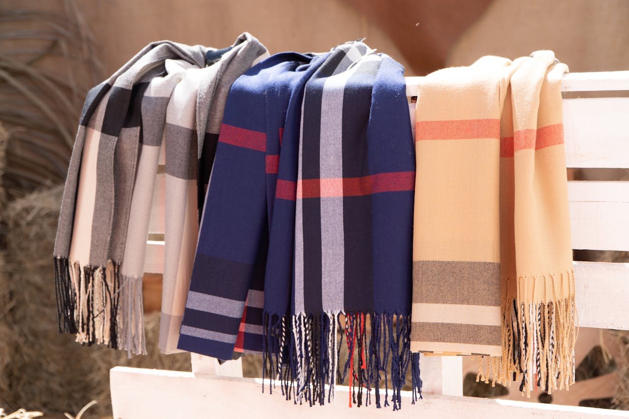 services-02 A variety of wool scarves draped over a white wooden rack in natural sunlight, showcasing vibrant colors and patterns.